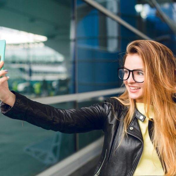 Close-up portrait of young girl standing outside in airport. She has long hair, black jacket and glasses. She is making selfie portrait.